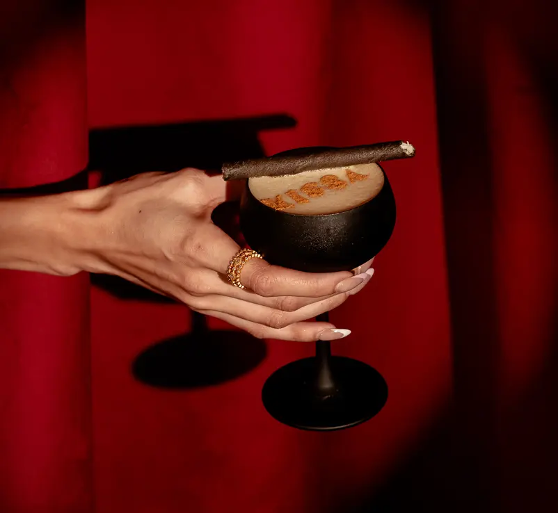 Hand holding a black espresso martini glass topped with creamy foam and a dark garnish against a dramatic red background.