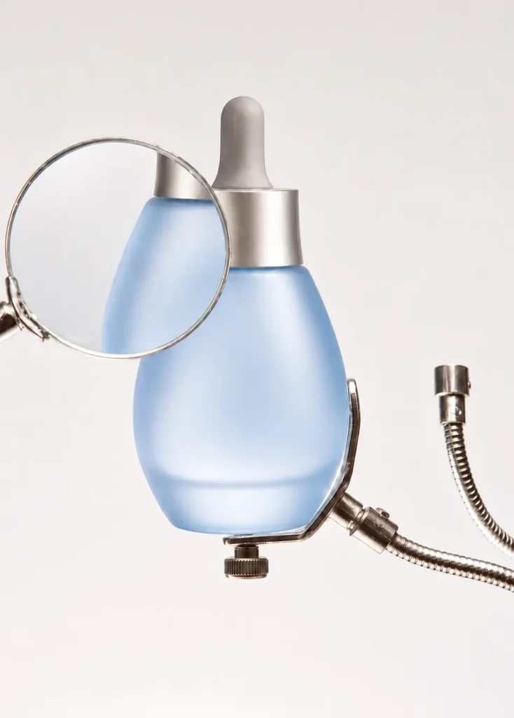 Frosted blue skincare serum bottle with a silver dropper, magnifying glass and metallic accessories against a white background.