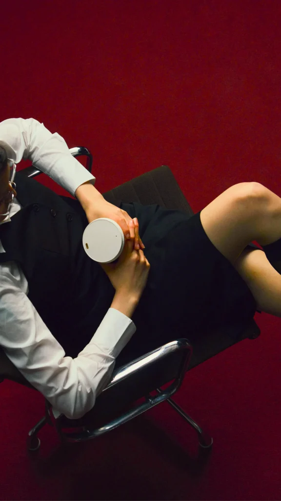 Fashion model reclining in a black chair, wearing a white shirt and black outfit while holding a white takeaway cup against a deep red background.