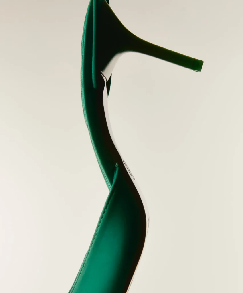 Close-up of two emerald green high-heeled shoes arranged in an abstract composition against a minimalist cream background.