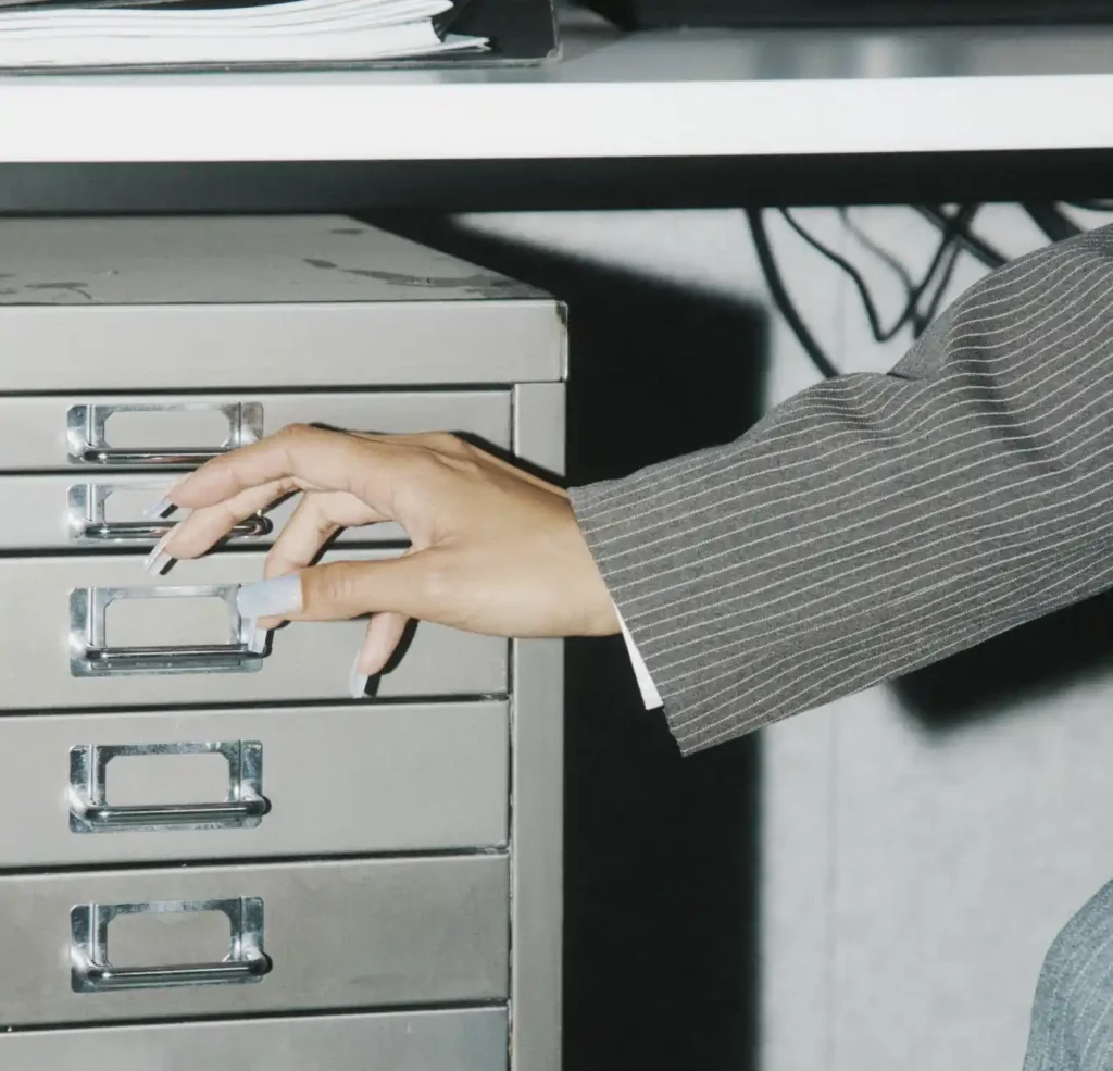 Close-up of a hand with long manicured nails reaching for a metal filing cabinet drawer while wearing a grey pinstripe blazer.