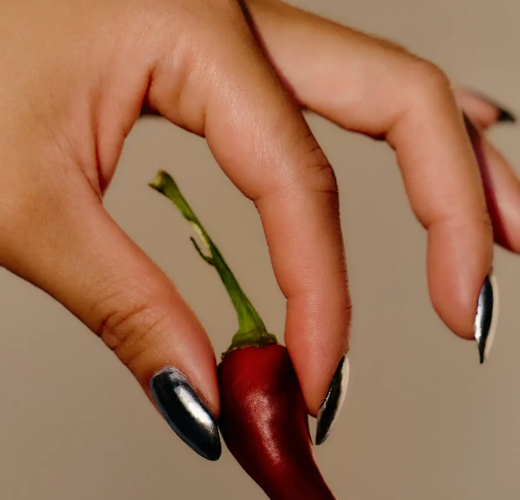 Close-up of a hand with metallic black nails delicately holding a red chilli pepper against a neutral background.