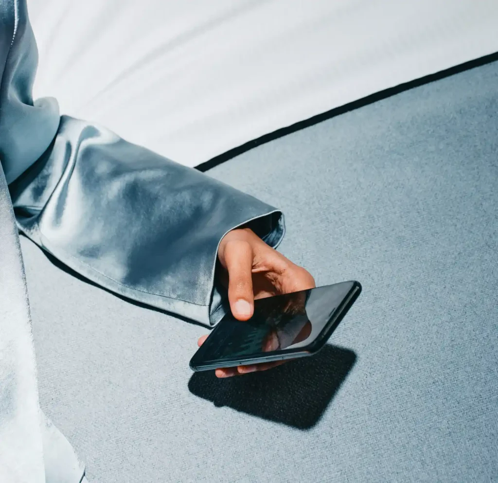 Close-up of a hand holding a black smartphone, emerging from a metallic blue sleeve against a textured grey background.