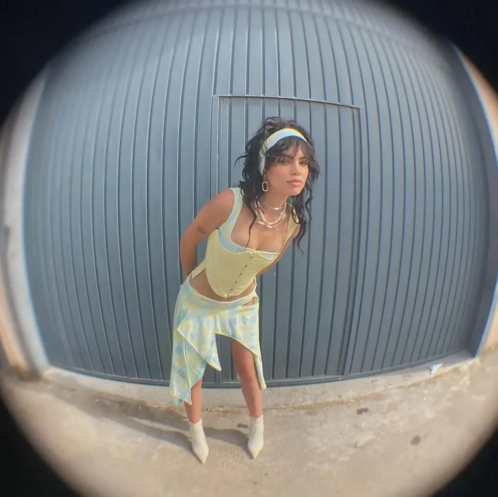 Fashion model wearing a pastel yellow and blue two-piece outfit with white boots, posing in front of a grey metal door.