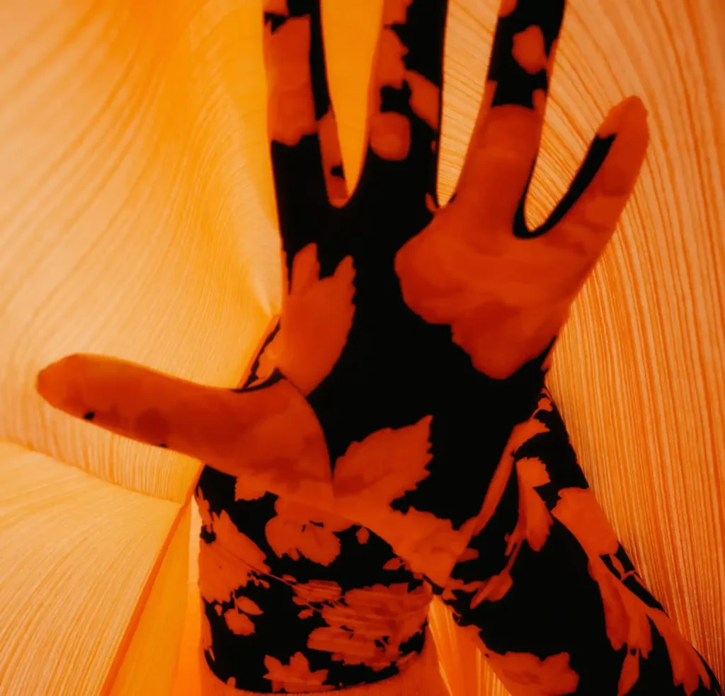 Close-up of outstretched hands and a black floral-patterned outfit illuminated by intense orange lighting against a textured background.