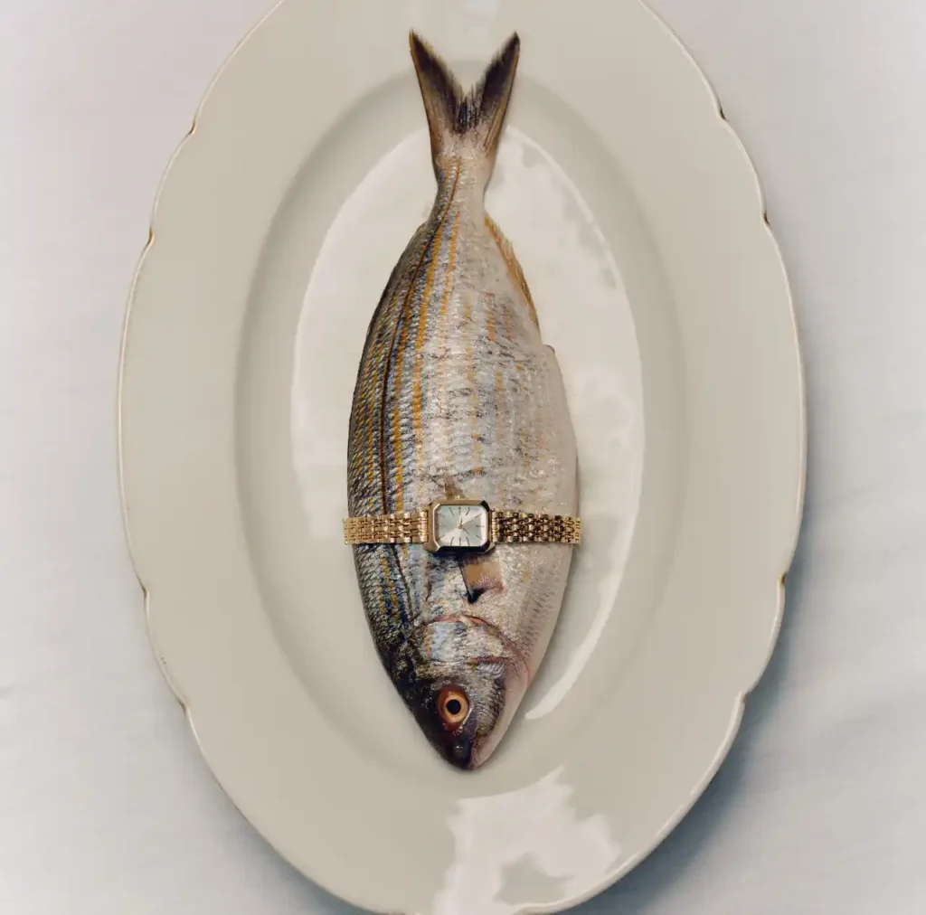 Silver striped fish presented on a white oval plate with a gold wristwatch wrapped around its body.