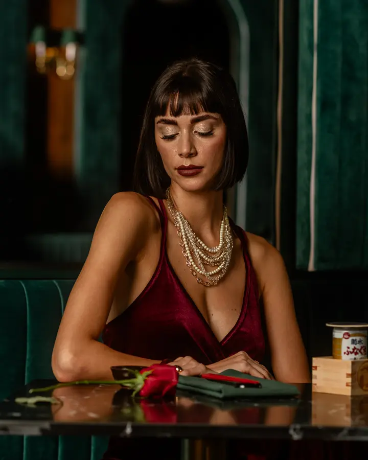 Fashion model with a short dark bob wearing a burgundy satin dress and layered pearl necklaces, seated at a table with a red rose.