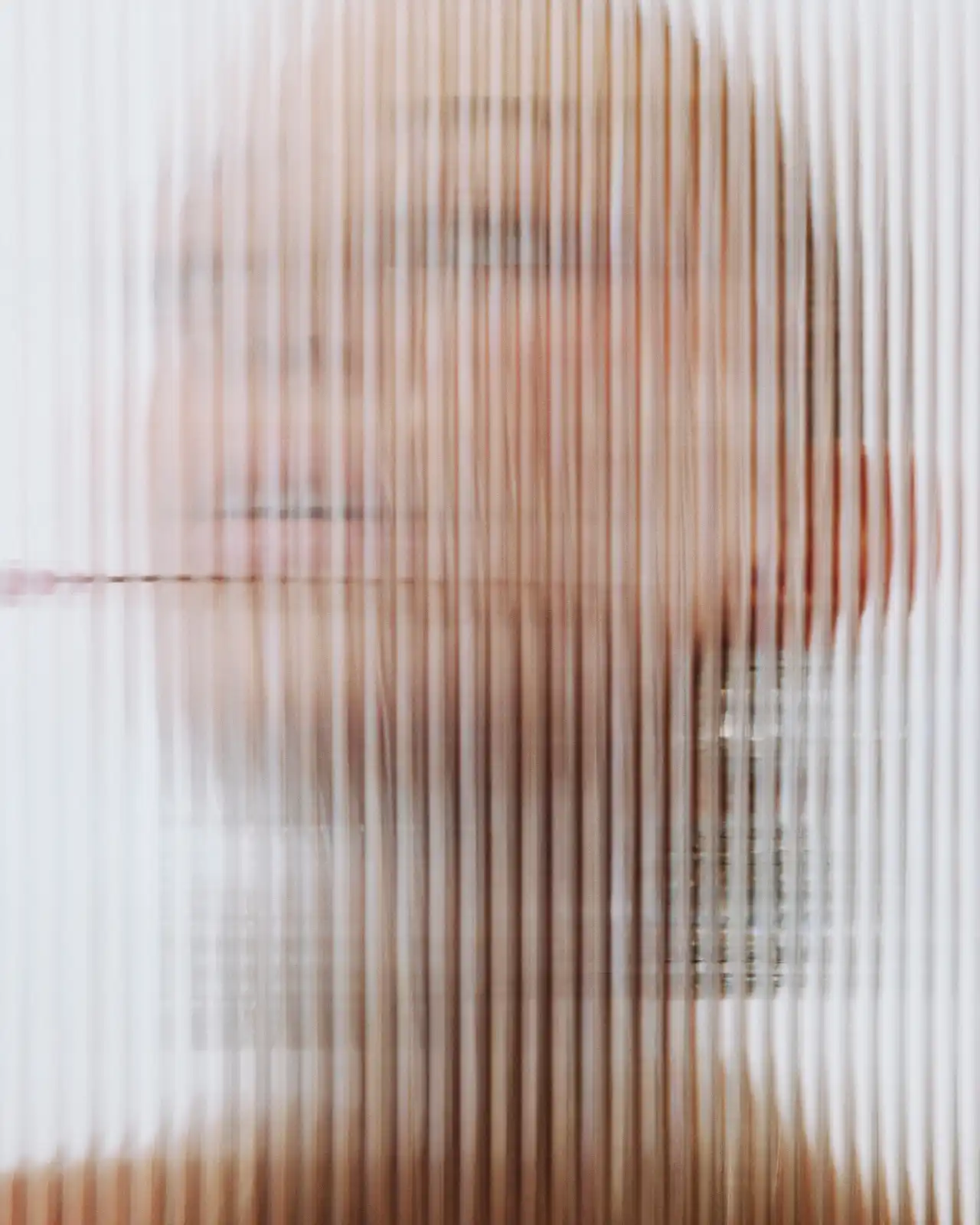 Abstract close-up portrait of a woman's face partially obscured by vertically ribbed translucent glass, creating a soft blurred effect.